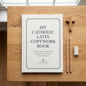 May include: A white hardback book titled My Catholic Latin Copywork Book with dark blue text, a wooden cross, two wooden pencils, and a small rectangular eraser. The cover features a black border and a small cross icon at the bottom centre.