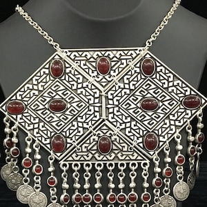 May include: A silver necklace with a large, geometric pendant featuring a repeating pattern and red gemstones. The pendant has a fringe of silver chains with red beads and coin-like charms.
