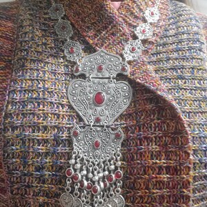 May include: A silver necklace with a large, ornate pendant featuring red stones. The pendant is shaped like a heart and has intricate details. The necklace is on a brown and blue patterned sweater.