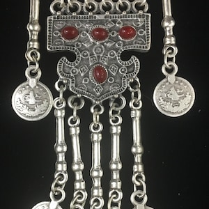 Peut inclure: Pendentif en argent avec une pierre rouge et des motifs complexes. Le pendentif a un sommet cylindrique avec une inscription en arabe et pend à une chaîne avec plusieurs breloques en forme de pièces de monnaie.