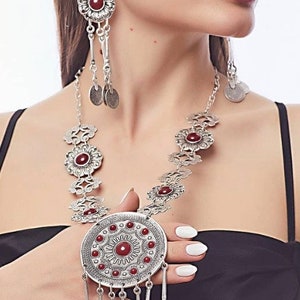 Peut inclure: Un collier en argent avec un grand pendentif rond orné d'une pierre rouge et de détails complexes. Le collier est orné de petits pendentifs circulaires avec des pierres rouges et une chaîne délicate. Des boucles d'oreilles assorties avec un design similaire complètent l'ensemble.