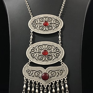 May include: A silver necklace with three oval pendants, each featuring intricate filigree designs and a red gemstone. The bottom pendant has a fringe of silver coins.
