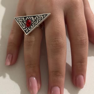 Puede incluir: Un anillo de plata con un diseño triangular y una piedra roja en el centro.