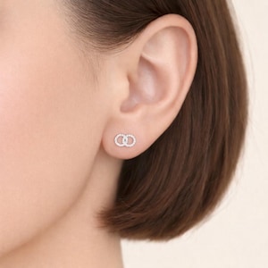 Puede incluir: Primer plano de una oreja con un pequeño pendiente de oro blanco. El pendiente presenta un diseño de doble círculo, incrustado con pequeños cristales brillantes. El pendiente está en la oreja.