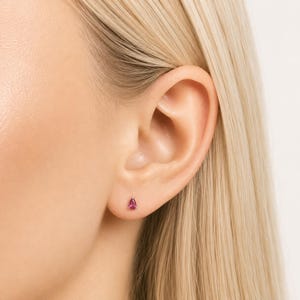 Può includere: Primo piano di un orecchio con un piccolo orecchino a bottone a forma di goccia, color rubino. L'orecchino è incastonato in un design semplice. La persona ha i capelli biondi.