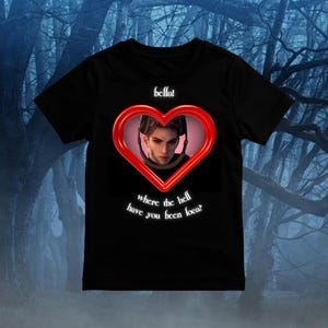 Puede incluir: Una camiseta negra de manga corta con un marco rojo en forma de coraz&oacute;n que contiene la ilustraci&oacute;n de una persona de pelo corto con auriculares. Sobre el coraz&oacute;n aparece el texto "bella!" y debajo "where the hell have you been loca?"