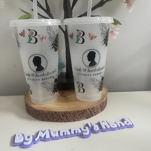 May include: Two clear plastic tumblers with lids and straws, decorated with butterflies, bees, and the text "Lady Whistledown's Society Papers." The tumblers are on a wooden coaster, with a purple sign that reads "By Mummy's Hand."