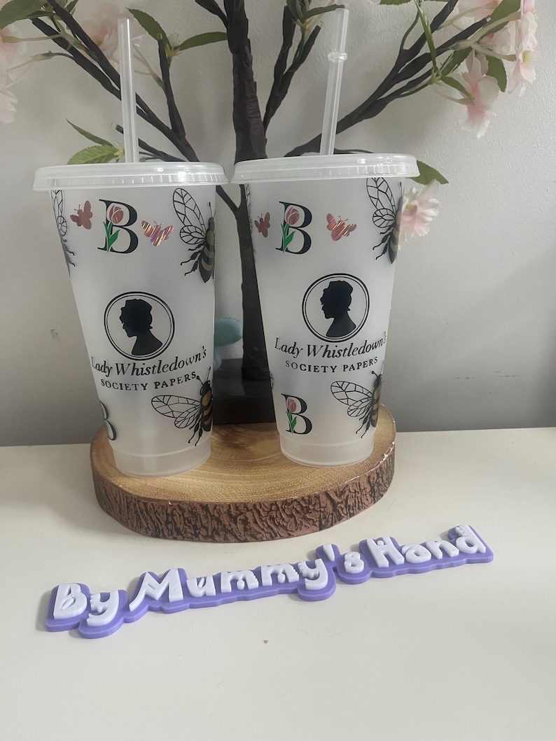 May include: Two clear plastic tumblers with lids and straws, decorated with butterflies, bees, and the text "Lady Whistledown's Society Papers." The tumblers are on a wooden coaster, with a purple sign that reads "By Mummy's Hand."