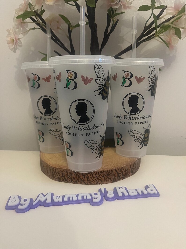 May include: Three clear plastic tumblers with lids and straws, decorated with a bee, butterfly, and the letter "B". Each tumbler features a silhouette and the text "Lady Whistledown's Society Papers". The tumblers are displayed on a wooden slice.