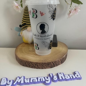 May include: A clear plastic tumbler with a lid and straw, decorated with bees, butterflies, and the text "Lady Whistledown's Society Papers." The tumbler also features the letter "B" and a silhouette of a woman. A gnome and a wooden slice are in the background.