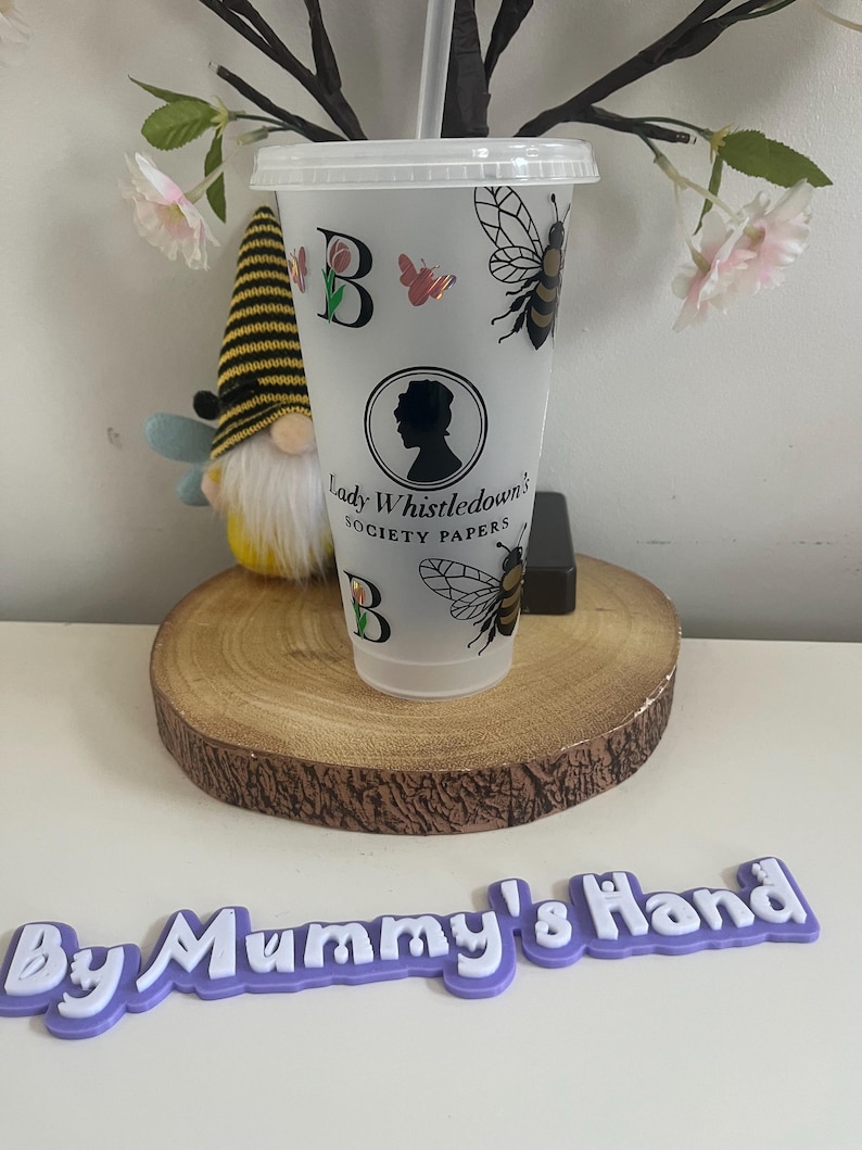 May include: A clear plastic tumbler with a lid and straw, decorated with bees, butterflies, and the text "Lady Whistledown's Society Papers." The tumbler also features the letter "B" and a silhouette of a woman. A gnome and a wooden slice are in the background.