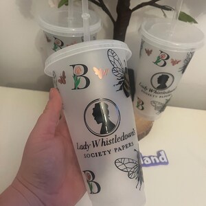May include: Frosted plastic tumbler with a lid and straw. The tumbler features a black and gold bee, butterfly, and rose design, along with the text "Lady Whistledown's Society Papers". The letter "B" is also featured on the tumbler.