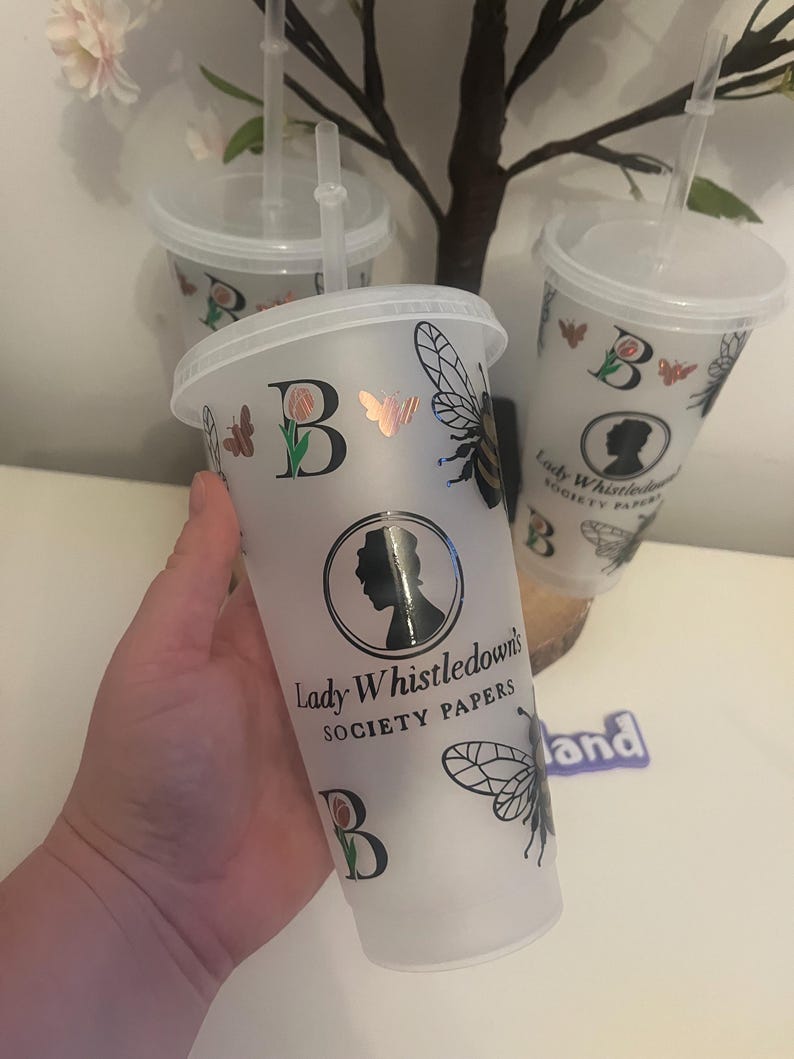 May include: Frosted plastic tumbler with a lid and straw. The tumbler features a black and gold bee, butterfly, and rose design, along with the text "Lady Whistledown's Society Papers". The letter "B" is also featured on the tumbler.