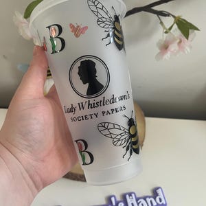 May include: A clear plastic tumbler with a white lid, featuring a black and gold bee, a butterfly, and the letter "B" with a tulip. The tumbler has the text "Lady Whistledown's Society Papers" and a silhouette of a woman.
