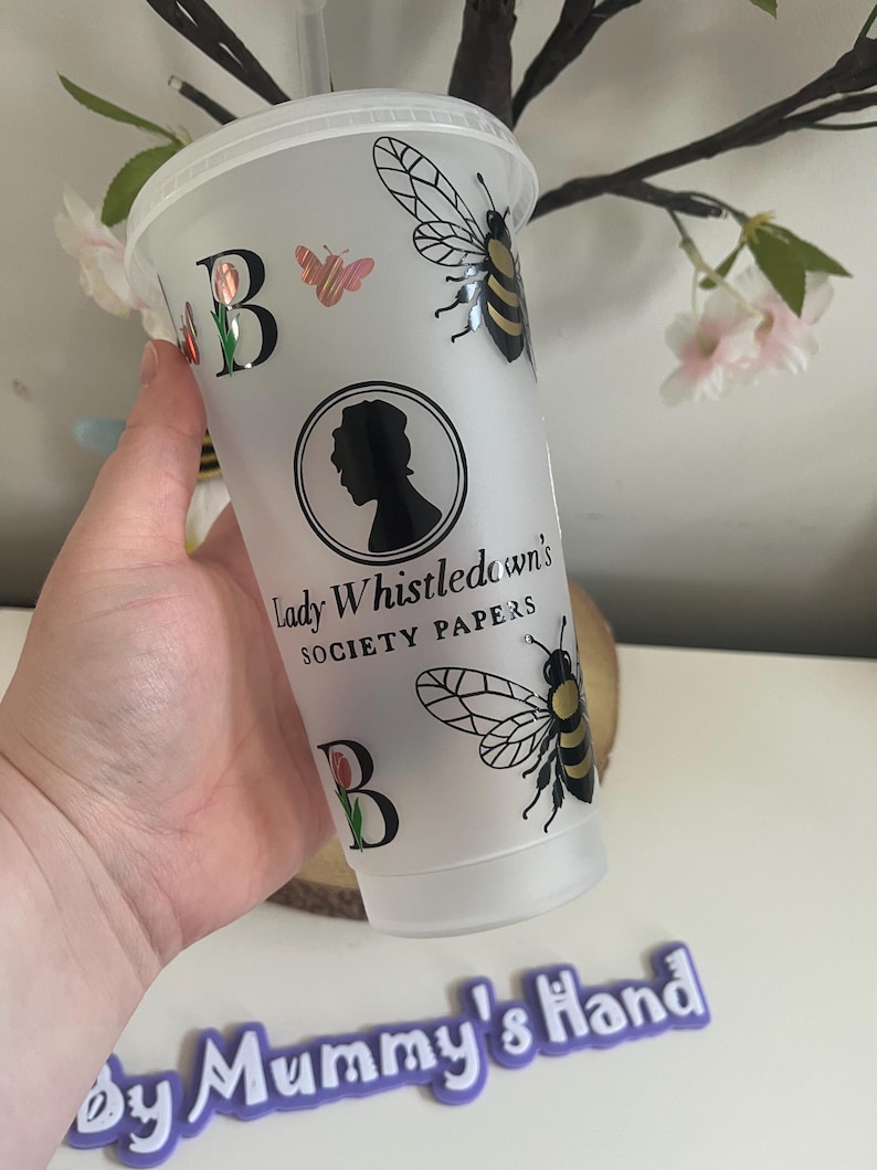 May include: A clear plastic tumbler with a white lid, featuring a black and gold bee, a butterfly, and the letter "B" with a tulip. The tumbler has the text "Lady Whistledown's Society Papers" and a silhouette of a woman.