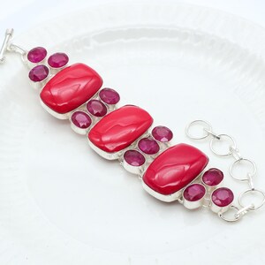 Può includere: Un bracciale in argento con grandi pietre rosse ovali e pietre rosa scuro rotonde più piccole. Il bracciale ha una chiusura a toggle ed è posto su una superficie bianca. Un accessorio audace.