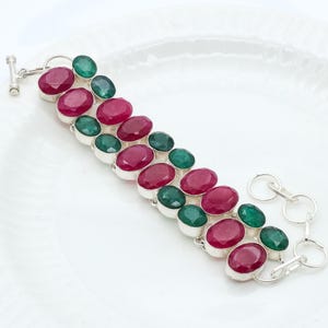 Puede incluir: Una pulsera de plata con filas alternas de piedras preciosas ovaladas de color rojo intenso y verde esmeralda. La pulsera tiene un cierre de palanca y se muestra sobre una superficie blanca. Las piedras están engastadas en biseles individuales de plata.