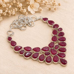 May include: A silver necklace featuring multiple teardrop-shaped, deep red ruby gemstones arranged in a layered, cascading pattern. The piece includes a silver chain with circular links and a toggle clasp closure, showcasing a polished metallic finish.