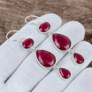 May include: A pair of drop earrings featuring faceted, deep red ruby gemstones set in silver-toned metal. Each earring consists of three stones: a small round top stone, a larger pear-shaped middle stone, and a small pear-shaped bottom drop.