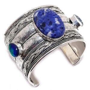Pode incluir: Uma pulseira de punho larga de prata com três pedras preciosas. A maior é uma sodalita azul oval, ladeada por uma pedra azul-esverdeada menor e uma pedra azul clara. A pulseira tem detalhes trançados e gravados.