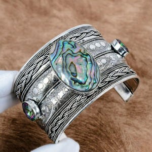 May include: Silver-toned cuff bracelet featuring an oval abalone shell centerpiece with iridescent colors. The bracelet has intricate braided and dotted patterns, and two smaller, colorful gemstone accents.