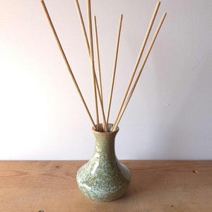 May include: A small, bulbous ceramic vase with a glossy, mottled sage green and white speckled glaze. The vase has a narrow neck and contains several thin, straight, light-coloured wooden reed diffuser sticks fanned out from the opening.