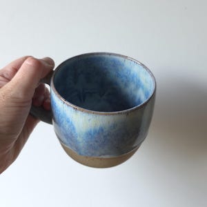 May include: A ceramic mug featuring a rounded shape with a handle. The upper portion of the mug has a glossy glaze in shades of blue and white, while the lower portion is finished in a matte, unglazed tan colour with a visible texture.