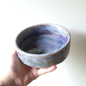 May include: A round ceramic bowl featuring a variegated glaze with blended tones of deep purple, soft blue, and muted grey. The surface has a smooth, glossy finish with subtle colour transitions throughout the interior and exterior walls.
