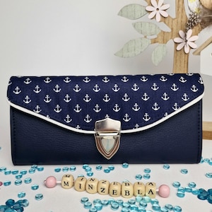 Könnte beinhalten: Eine dunkelblaue Brieftasche mit einem weißen Ankermuster und einem silbernen Verschluss. Die Brieftasche ist geschlossen und liegt auf einer weißen Oberfläche mit blauen und rosa Perlen.