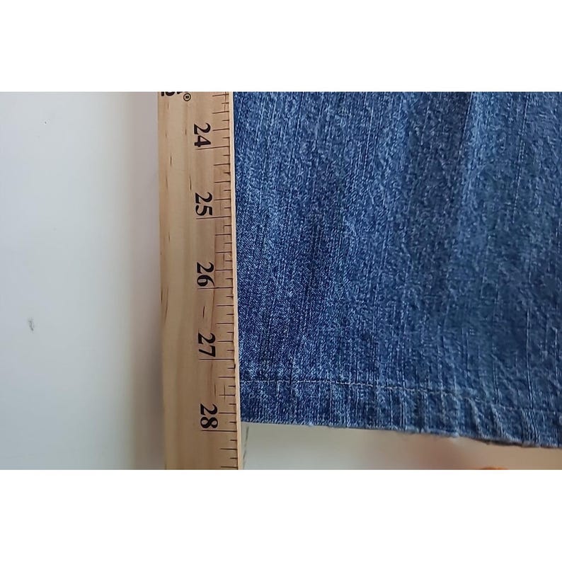 May include: A blue denim fabric section positioned next to a wooden ruler. The ruler shows measurements in inches, with the fabric edge aligning near the 24-inch mark and extending past the 28-inch mark, indicating a length of over 4 inches.