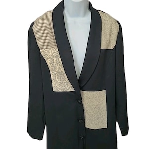 May include: A black blazer featuring a shawl collar and three front buttons. The garment includes contrasting beige and black patterned fabric panels on the upper chest and lower front, showcasing houndstooth and intricate floral designs.