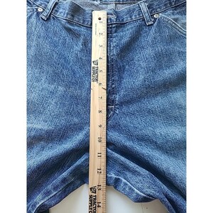 May include: Blue denim jeans featuring a classic five-pocket design with a button fly and belt loops. A wooden ruler is placed vertically over the center, showing a measurement of approximately 14 inches from the waistband down the front seam.