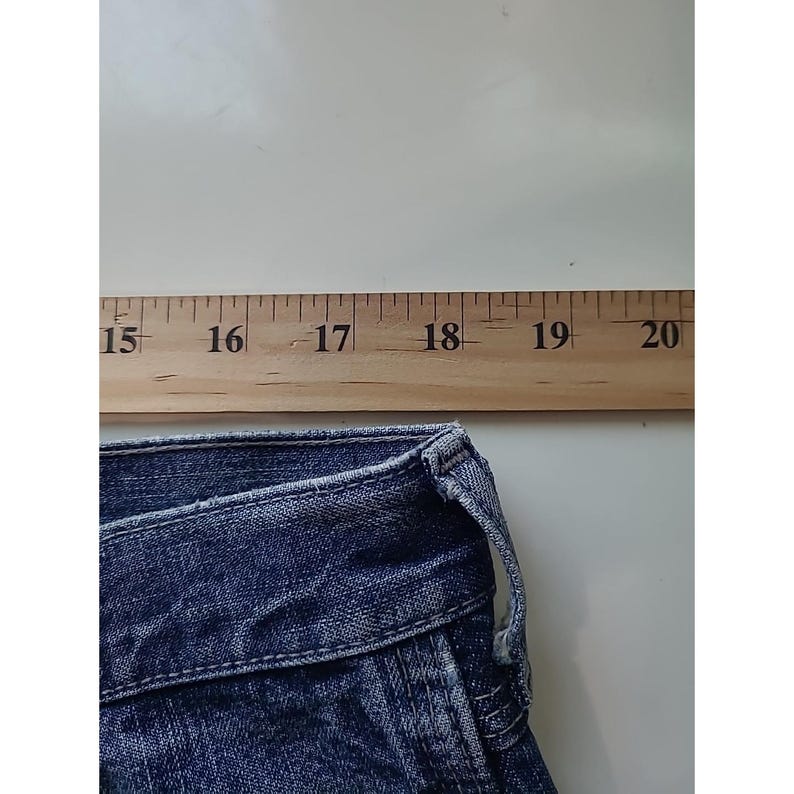 May include: A pair of blue denim jeans featuring a waistband with a belt loop, positioned against a wooden ruler that shows a measurement of approximately 5 inches across the top edge of the garment, highlighting the texture of the fabric.