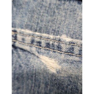 May include: A close-up view of light blue denim fabric featuring a horizontal seam with double-stitched light-colored thread. The material displays a distressed texture with visible fraying and loose white threads along the stitched edge.