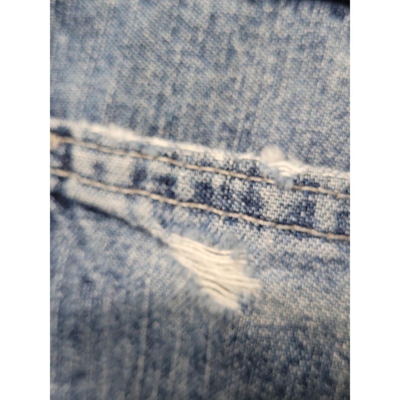 May include: A close-up view of light blue denim fabric featuring a horizontal seam with double-stitched light-colored thread. The material displays a distressed texture with visible fraying and loose white threads along the stitched edge.