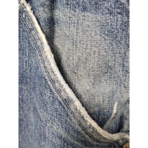 May include: A close-up view of blue denim fabric featuring a curved pocket opening with a frayed, distressed white edge. The material displays a classic medium-wash blue texture with visible woven threads and a subtle worn-in appearance throughout.