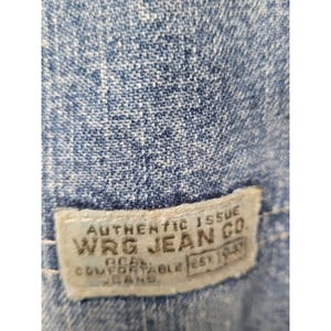 May include: A blue denim fabric with a rectangular off-white fabric label stitched onto it. The label features the text AUTHENTIC ISSUE WRG JEAN CO. REAL COMFORTABLE JEANS EST 1947 in dark lettering. The denim has a visible woven texture.