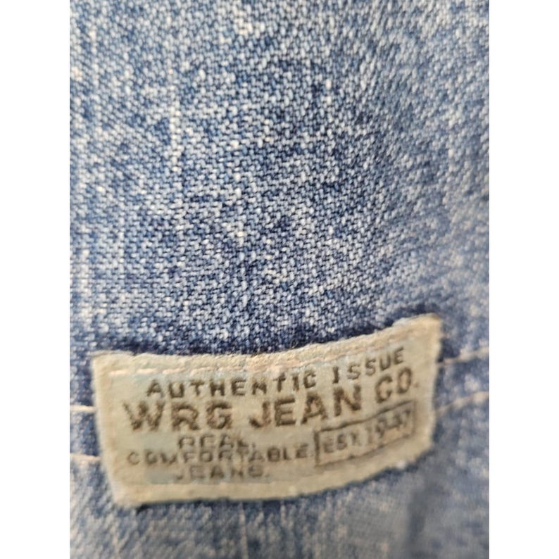 May include: A blue denim fabric with a rectangular off-white fabric label stitched onto it. The label features the text AUTHENTIC ISSUE WRG JEAN CO. REAL COMFORTABLE JEANS EST 1947 in dark lettering. The denim has a visible woven texture.