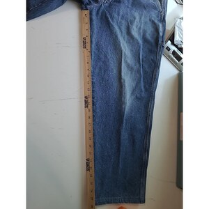 May include: A pair of blue denim jeans featuring a faded wash and visible stitching along the side seams. A wooden ruler is placed vertically along the side of the leg, showing a length measurement extending from zero to twenty-eight inches.