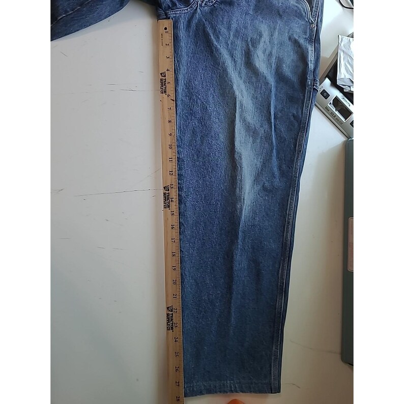 May include: A pair of blue denim jeans featuring a faded wash and visible stitching along the side seams. A wooden ruler is placed vertically along the side of the leg, showing a length measurement extending from zero to twenty-eight inches.