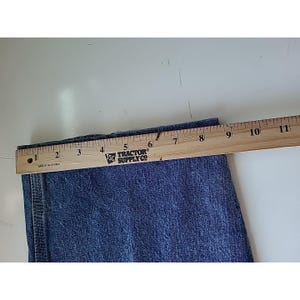 May include: A blue denim pant leg featuring a vertical side seam and a hemmed edge, positioned beneath a wooden ruler that shows a width measurement of approximately 11 inches across the fabric from the left edge to the right side.