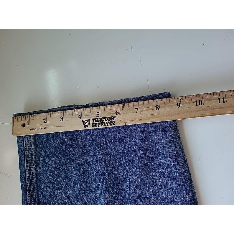 May include: A blue denim pant leg featuring a vertical side seam and a hemmed edge, positioned beneath a wooden ruler that shows a width measurement of approximately 11 inches across the fabric from the left edge to the right side.