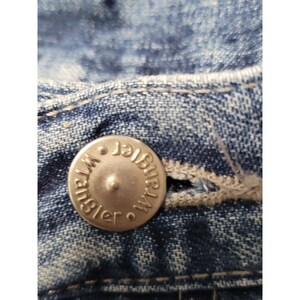 May include: A close-up view of a blue denim fabric featuring a silver-toned metal button embossed with the word Wrangler. The button is centered on the waistband, which includes a stitched buttonhole and visible light-colored thread detailing.