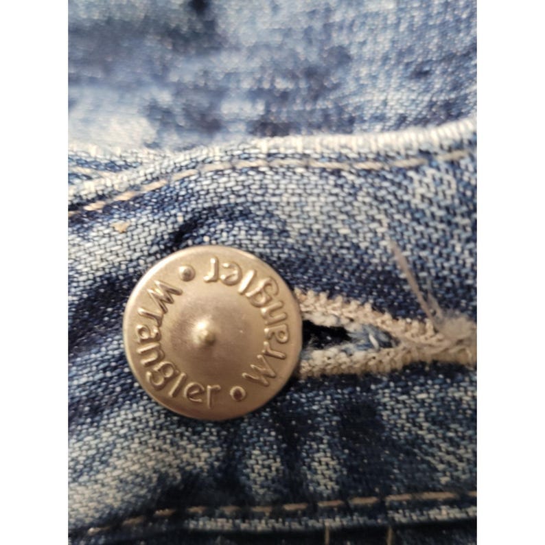 May include: A close-up view of a blue denim fabric featuring a silver-toned metal button embossed with the word Wrangler. The button is centered on the waistband, which includes a stitched buttonhole and visible light-colored thread detailing.