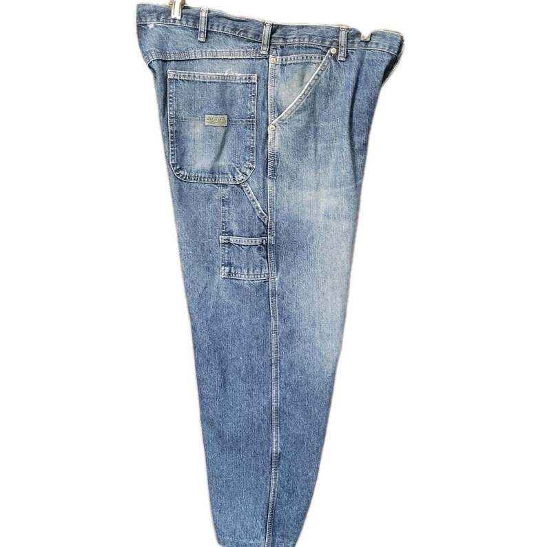 May include: Light blue denim carpenter pants featuring a high-waisted design, a metal button closure, belt loops, and multiple utility pockets on the side. The fabric displays a faded wash with visible stitching and a rectangular brand patch.