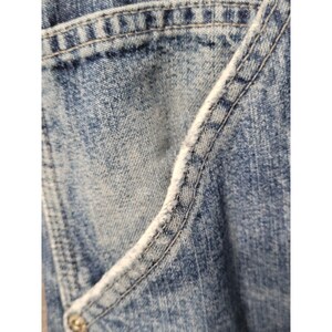 May include: A close-up view of blue denim fabric featuring a light acid-wash finish. The material displays a diagonal pocket opening with visible frayed, white distressed edges along the seam and reinforced stitching throughout the garment.