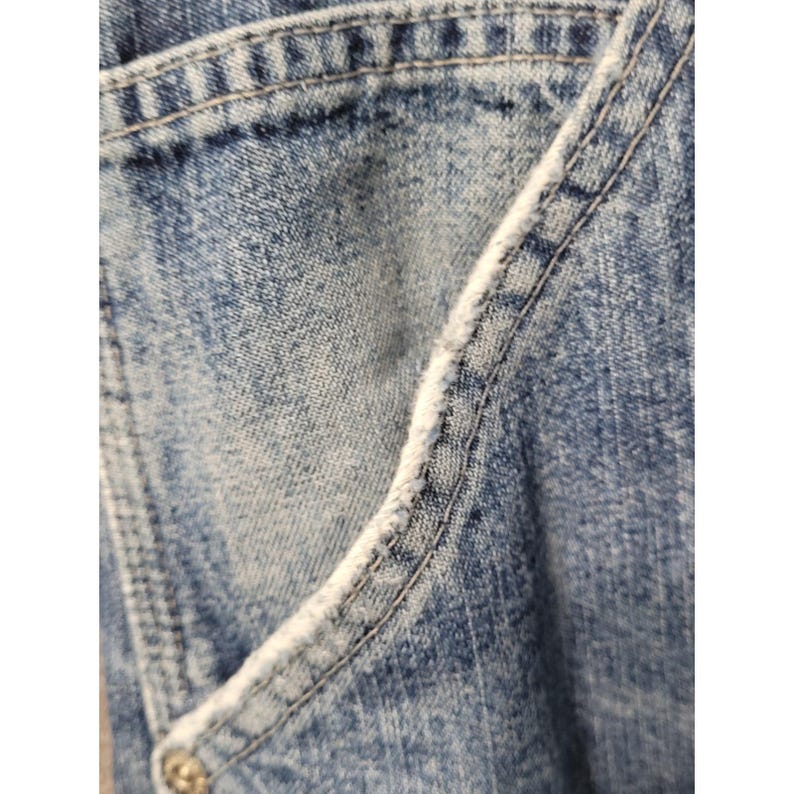 May include: A close-up view of blue denim fabric featuring a light acid-wash finish. The material displays a diagonal pocket opening with visible frayed, white distressed edges along the seam and reinforced stitching throughout the garment.
