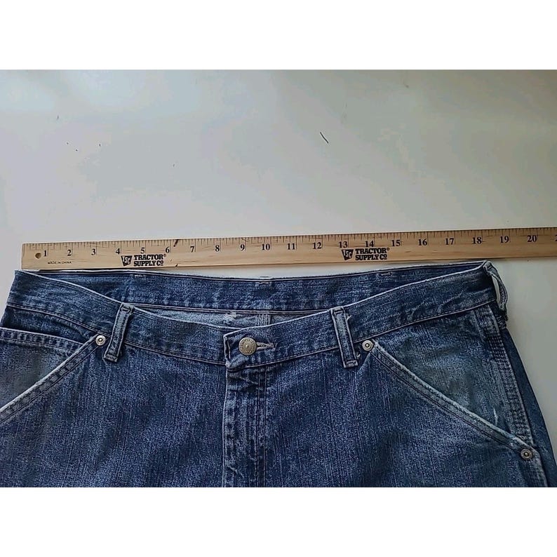 May include: Blue denim jeans featuring a button fly closure, belt loops, and front pockets. A wooden ruler is positioned along the top waistband, showing a measurement of approximately 20 inches across the width of the waist area.