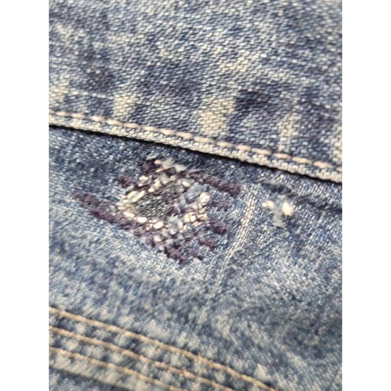 May include: A close-up view of blue denim fabric featuring a distressed, acid-wash texture with a visible horizontal seam. A small, square-shaped area of dark blue thread repair or darning is present on the surface, showing dense stitching.
