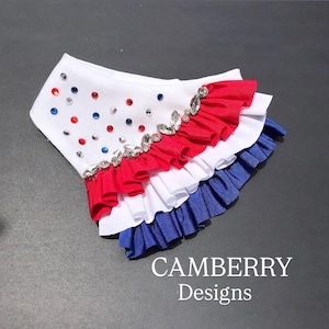 May include: A white pet bandana featuring red and blue rhinestones scattered across the top section. The lower portion consists of three layered fabric ruffles in red, white, and blue. The text CAMBERRY Designs is printed in white at the bottom.